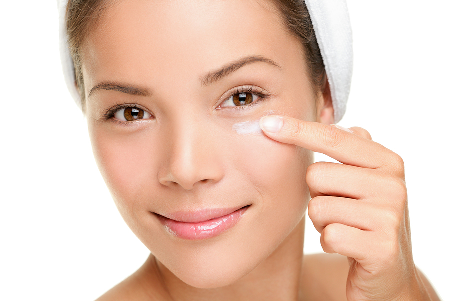 Crema facial mujer aplicando crema para la piel debajo de los ojos.  Crema de belleza para el contorno de ojos, crema antiarrugas o crema antiedad para el cuidado de la piel.  Hermosa joven mestiza asiática china / modelo de belleza femenina caucásica de unos 20 años aislada de fondo blanco.